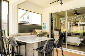 A modern outdoor kitchen area with a dining table and chairs, a built-in grill, and a sliding glass door leading to a patio. - Windsor Hills Luxe 4BR Villa w/ Pool & Game Room (Kissimmee)