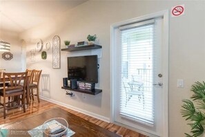 A living room with a TV on a wall-mounted shelf, a dining area with wooden chairs, and a sliding glass door with blinds. - Stylish 3BR Apartment at Vista Cay Resort (Orlando)