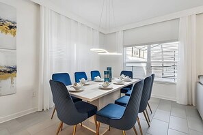 A modern dining room with a white table set for six, blue chairs, and a large window with blinds. - 5BR Storey Lake Townhome, Heated Pool (Kissimmee)