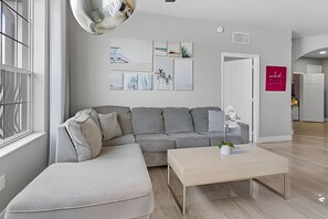 A modern living room with a grey sectional sofa, a small white ottoman, a wooden coffee table, and a potted orchid. There are abstract wall art pieces and a door with a sign on the wall. - Vista Cay 3BR Condo w/ Game Room & Balcony (Orlando)