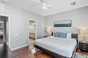 A bedroom with a large bed, a ceiling fan, a painting of the ocean, and a bathroom visible through an open door. - Elegant 3-bedroom Townhome at Vista Cay (Orlando)