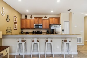A modern kitchen with wooden cabinets, a central island with bar stools, stainless steel appliances, and decorative wall art. - 4BR Paradise Palms Townhome With Pool (Kissimmee)