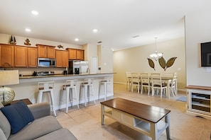 A modern kitchen with wooden cabinets, a central island with bar stools, a dining area with a chandelier, and a living room with a sofa and coffee table. - 4BR Paradise Palms Townhome With Pool (Kissimmee)