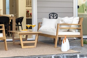 A wooden bench with cushions, a table with a glass and a plate, and a white bag on a mat outside a house. - Sunset View Updated Villa Heated Pool  Kings (Waikoloa Village)