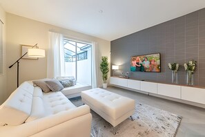Un salon moderne avec un canapé blanc, une table basse, un lampadaire, un téléviseur fixé au mur et une grande fenêtre avec des rideaux.