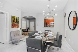 A modern dining area with a glass-top table set for six, surrounded by grey upholstered chairs. - Spacious 3BR Condo at Vista Cay Resort (Orlando)