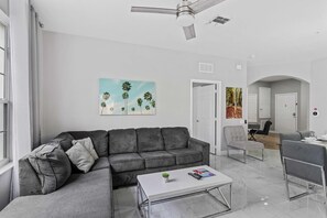 Ein modernes Wohnzimmer mit einer grauen Eckcouch, einem weißen Couchtisch und einem Deckenventilator. Zwei Türen führen in andere Räume, und an der Wand hängt ein Palmenbild.