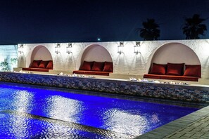 A poolside area with a blue pool, a seating area with red cushions, and wall-mounted lights. - Casa Frida | 7BR Spanish Estate w/ Pool, Spa & Cabanas (Coachella)