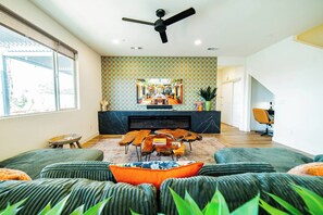 A modern living room with a fireplace, a ceiling fan, and a large window. - Casa Frida | 7BR Spanish Estate w/ Pool, Spa & Cabanas (Coachella)