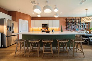 A modern kitchen with a central island, bar stools, a refrigerator, and a microwave. - Casa Frida | 7BR Spanish Estate w/ Pool, Spa & Cabanas (Coachella)