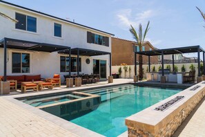 A swimming pool with a sunken seating area, a covered patio with outdoor furniture, and a building with large windows and a flat roof. - Casa Frida | 7BR Spanish Estate w/ Pool, Spa & Cabanas (Coachella)