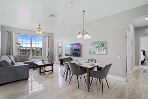 A modern living room with a sofa, dining table, and chairs. There is a painting on the wall and a ceiling fan. - Spacious 3BR Apartment at Vista Cay Resort (Orlando)
