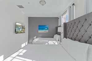 Une chambre moderne avec un grand lit, une tête de lit rembourrée de couleur grise, un téléviseur fixé au mur et un tableau représentant une scène de plage.