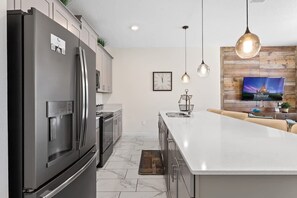 A modern kitchen with a large island, stainless steel appliances, pendant lighting, and a wooden accent wall with a TV. - Disney 4BR Townhome w/ Pool Storey Lake (Kissimmee)