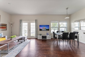 A modern living room with a dining area, wooden flooring, a flat-screen TV mounted on the wall, and a large window providing natural light. - 4BR West Lucaya Home Near Disney (Kissimmee)