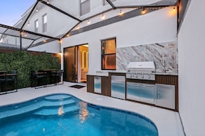 A modern outdoor kitchen with a built-in grill, a pool, and a seating area with chairs. - Modern 5BR Villa w/ Pool 15 min to Disney (Orlando)