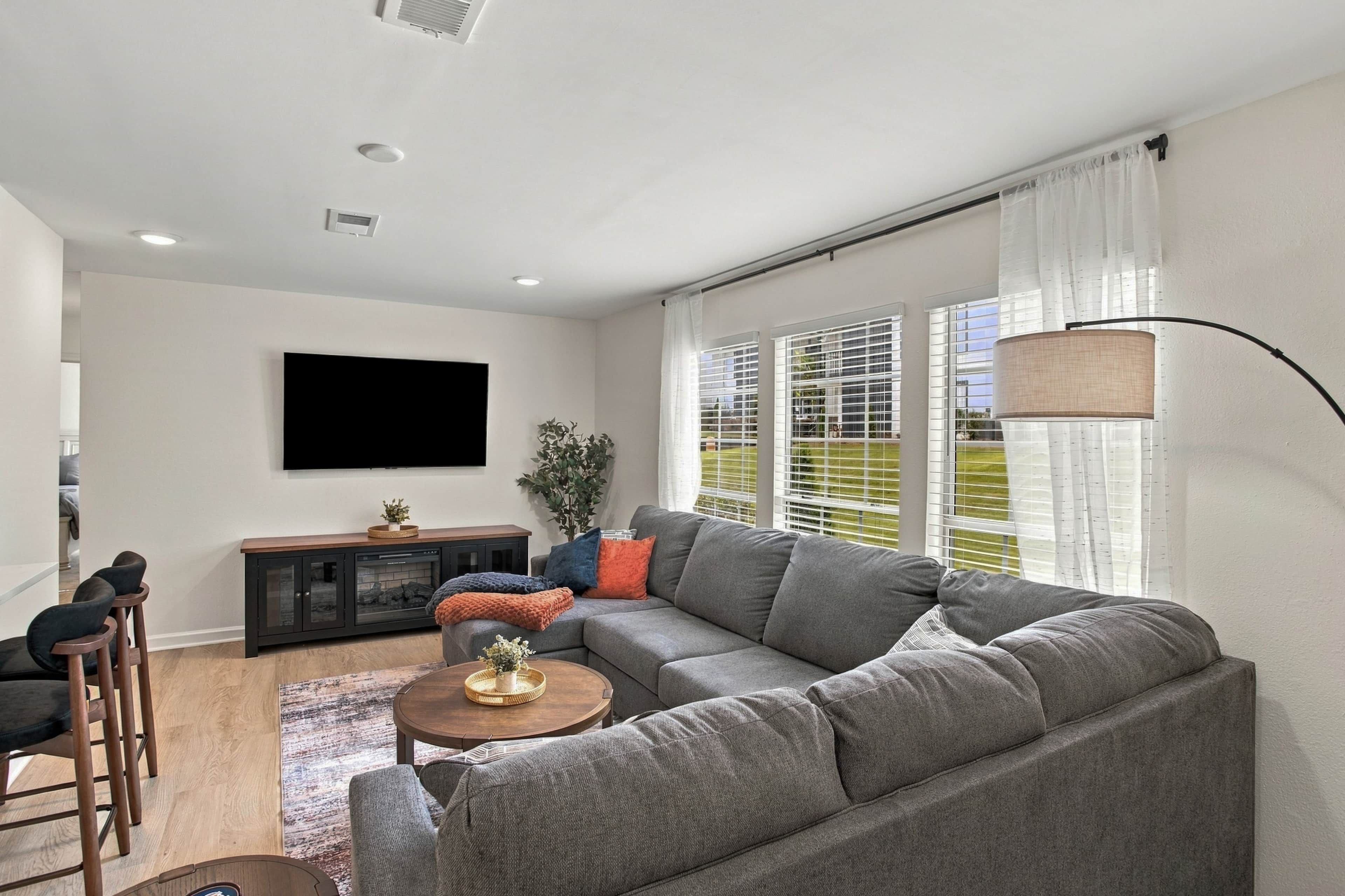 Ein modernes Wohnzimmer mit einem grauen Sofa, einem flachbildfernseher an der Wand, einem Holztisch und einer Stehlampe.
