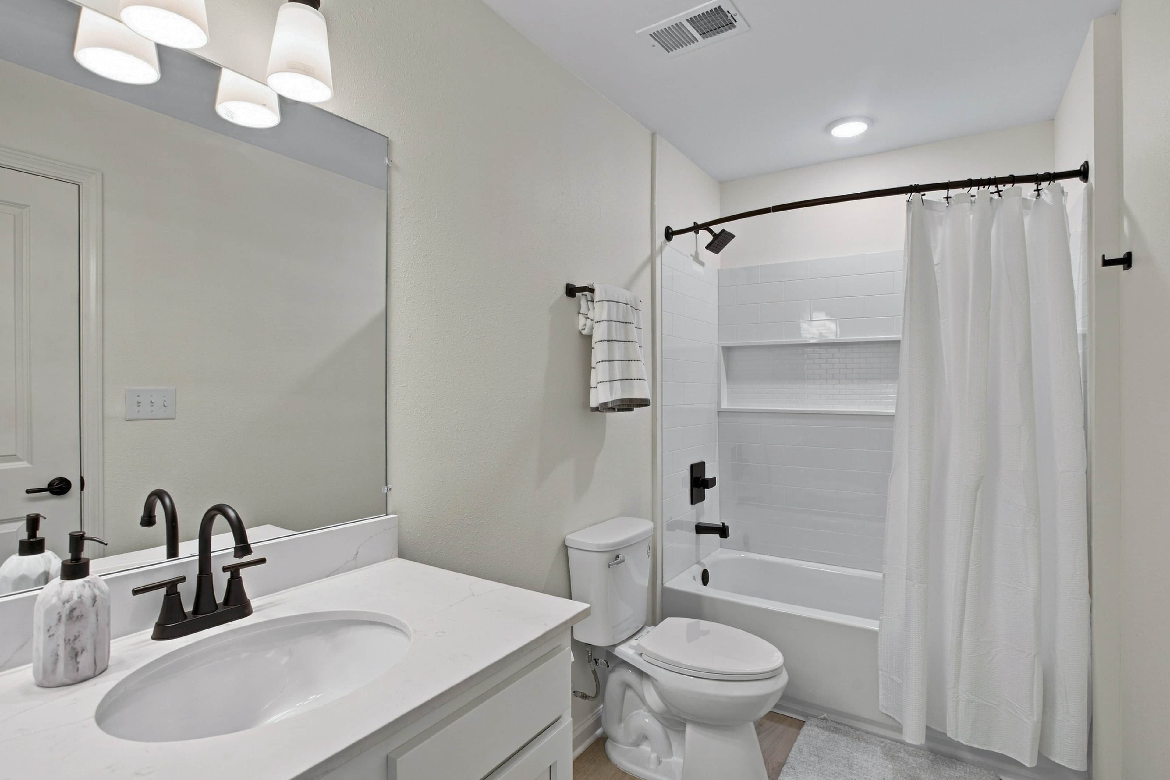 Un bagno moderno con lavandino bianco, rubinetto nero, water bianco e zona doccia con tenda bianca.