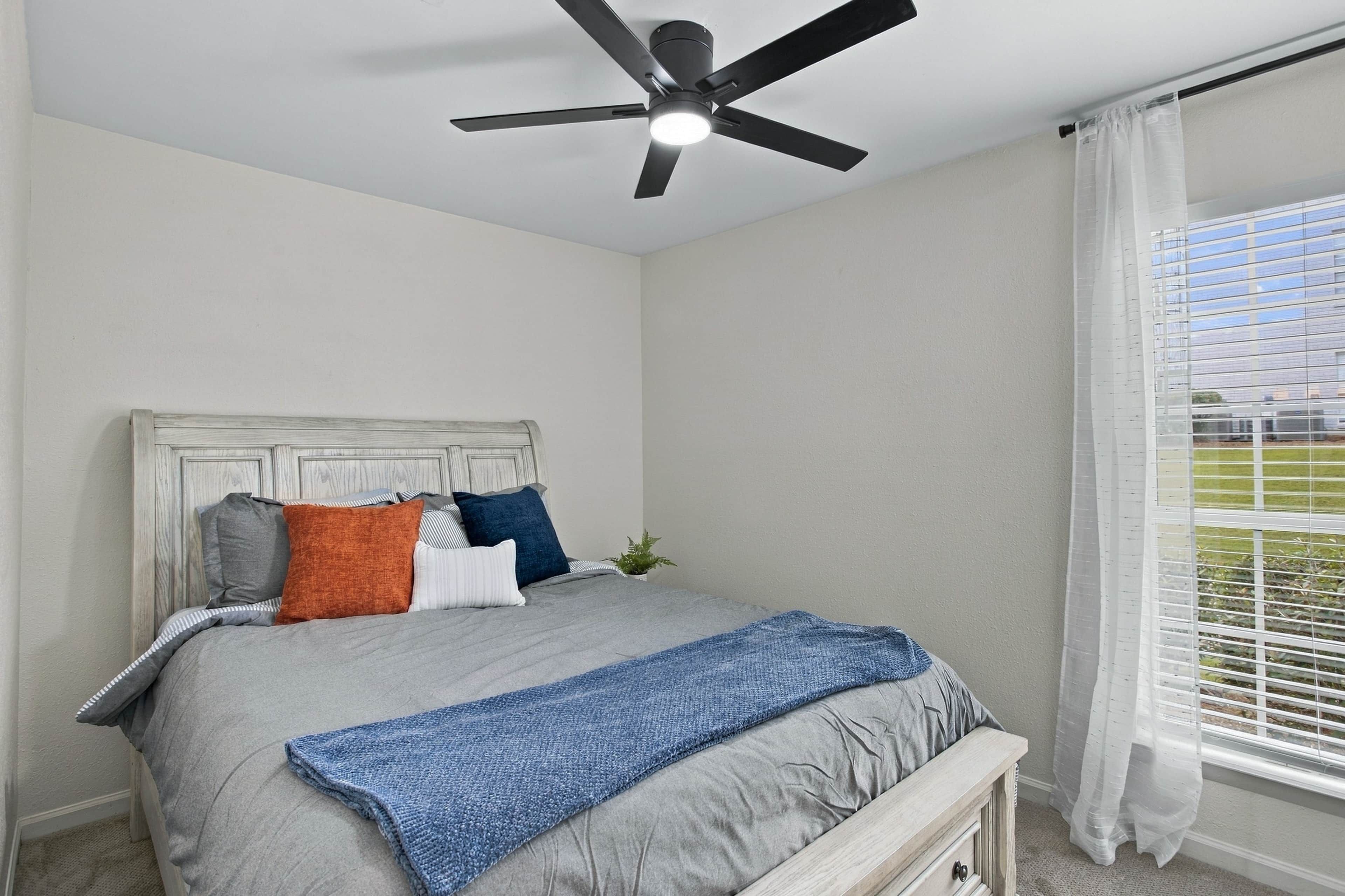 Ein Schlafzimmer mit einem Bett, einem Deckenventilator, einem Fenster mit Jalousien und Blick ins Freie.