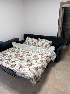 A bed with a patterned bedspread and two pillows, a black sofa, and a tiled floor.