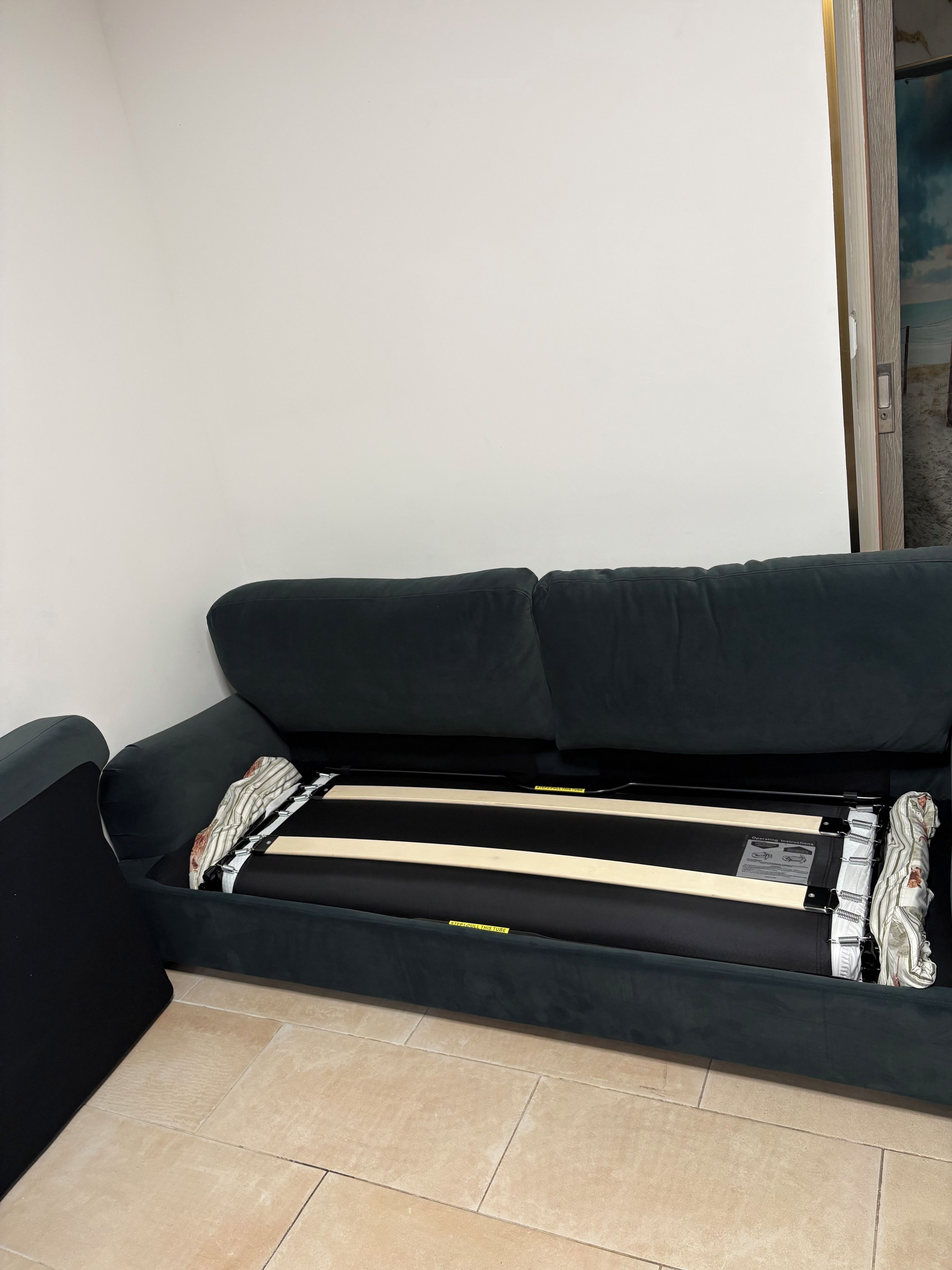 A black sofa with a mattress and pillows, a tiled floor, and a door leading to another room.