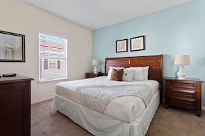 A bedroom with a large bed, two bedside tables, a dresser, a window with blinds, and two framed pictures on the wall. - Modern 4BR Home in Lucaya Village Kissimmee (Kissimmee)