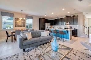 A modern living room with a grey sofa, a coffee table, and a dining area with chairs. - 5BR Solara Home w/ Private Pool Near Disney (Kissimmee)