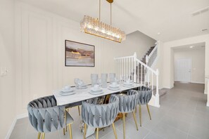 Un coin repas avec une longue table dressée pour six personnes, des chaises rembourrées, un lustre et un escalier en arrière-plan.