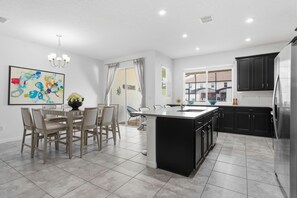 A modern kitchen with a central island, black cabinets, and a dining area with a table and chairs. - Solara 4BR w/ Private Pool Near Disney (Kissimmee)