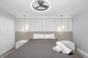 Ein modernes Schlafzimmer mit Bett, Nachttischen, Deckenventilator und einem Fenster mit Vorhängen.