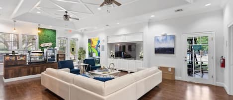 A spacious living room with a large sectional sofa, a kitchen area with a bakery sign, and a ceiling fan.