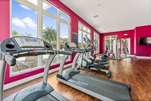 Ein Fitnessstudio mit Laufbändern, Crosstrainern und Krafttrainingsgeräten, großen Fenstern und einer roten Wand.