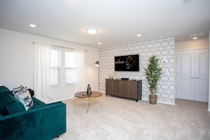 A living room with a teal sofa, a small round table, a TV on the wall, and a patterned wallpaper. - BR Villa w/ Pool, Star Wars Game Room + Resort (Davenport)