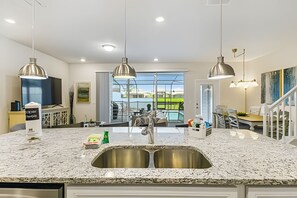 Une cuisine dotée d’un grand îlot central, d’un évier en acier inoxydable, de luminaires suspendus et offrant une vue sur une piscine grâce à de grandes fenêtres.