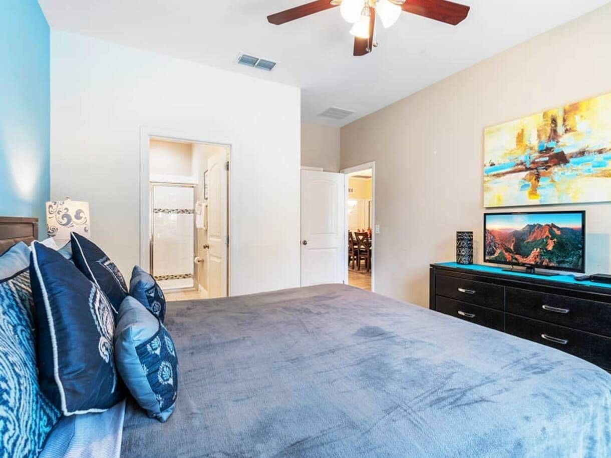 A bedroom with a bed, a ceiling fan, a TV, and a painting on the wall.