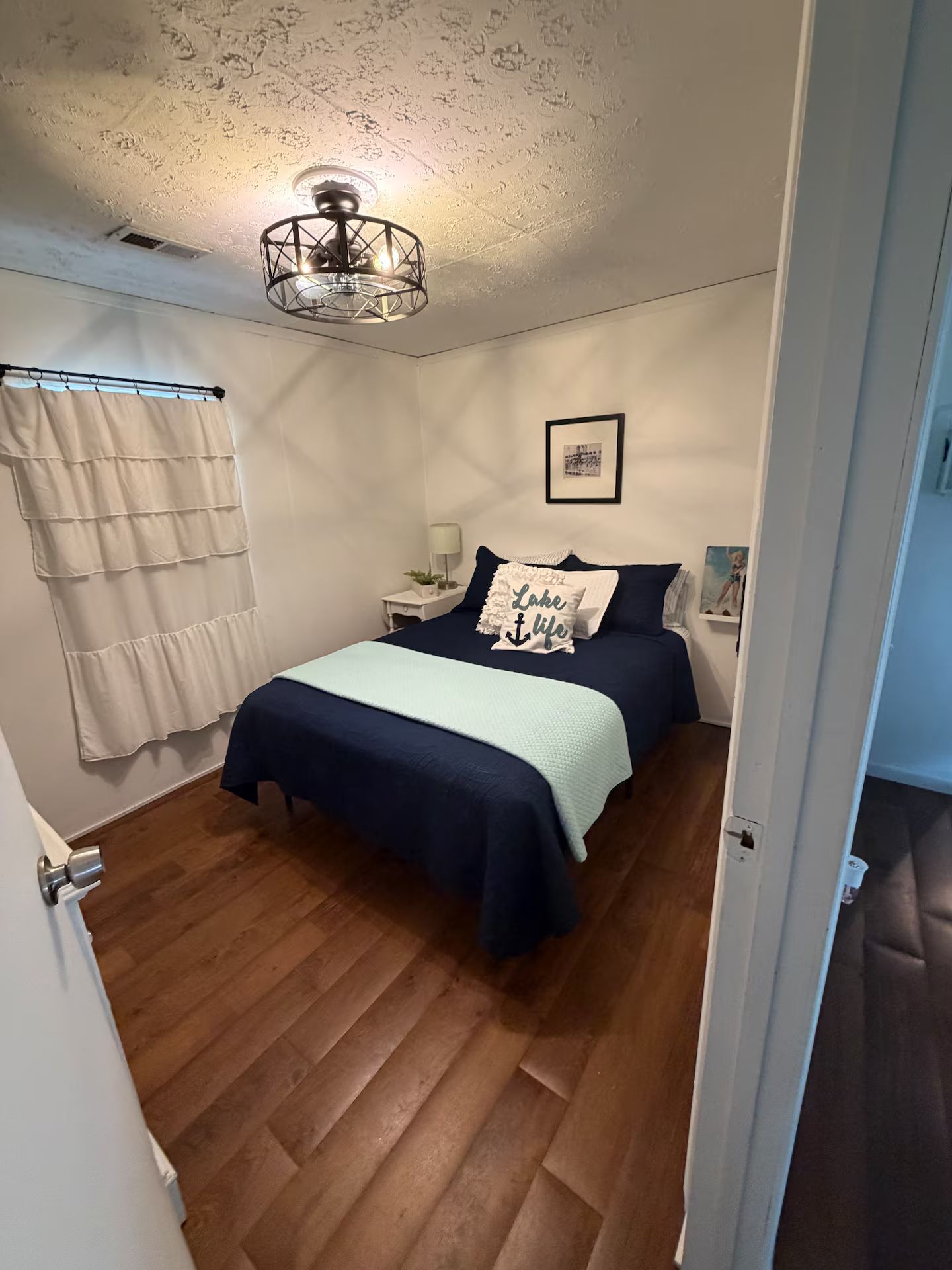 Un dormitorio con una cama, una mesita de noche, una lámpara, una lámpara del techo e imagen colgada en la pared.