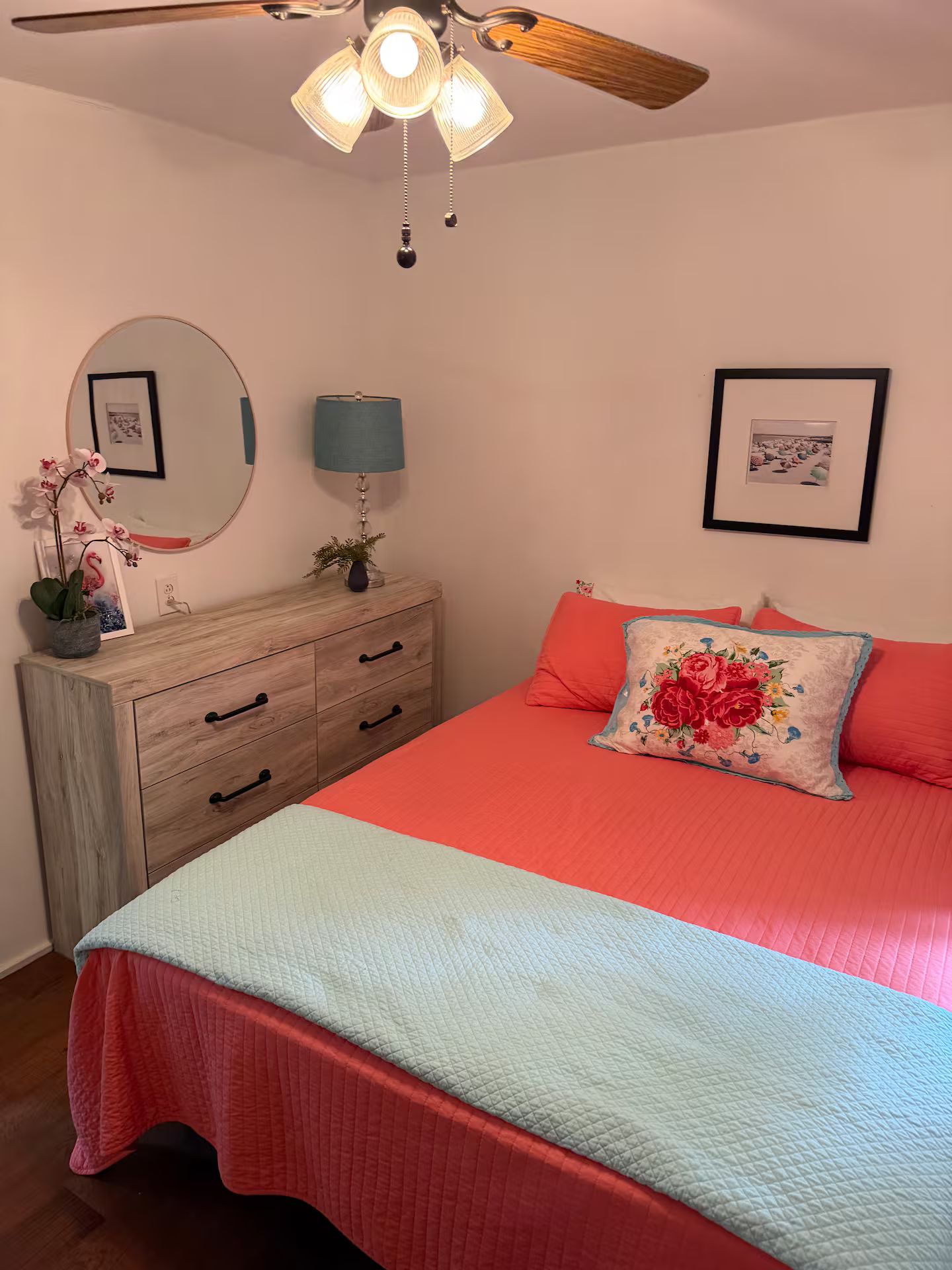 Une chambre à coucher comprenant un lit, une commode, une lampe, un ventilateur de plafond et deux tableaux encadrés accrochés au mur.