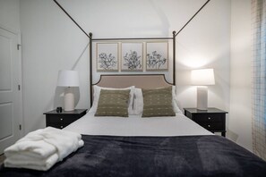 Un lit bien fait, recouvert d’une couverture foncée, avec deux oreillers et une serviette blanche pliée. On y trouve deux tables de chevet avec des lampes, et une œuvre d  ’art encadrée au-dessus du lit.