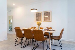 A dining area with a rectangular table set for six, surrounded by six brown upholstered chairs with metal frames. Above the table, there are two framed artworks on the wall. - Storey Lake 4BR Townhome - Pool & Themed Beds (Kissimmee)