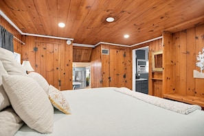 A bedroom with a wooden ceiling, a bed with white bedding, and a visible kitchen area with modern appliances. - Cozy Royal Pines Cottage w Courtyard & Patio (Royal Pines)