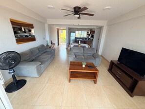 A living room with a sofa, a coffee table, a TV stand, and a ceiling fan. - Beautiful BEACHFRONT apartment for 4 people with a spectacular view. (Zihuatanejo)