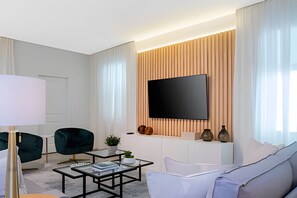 A modern living room with a flat-screen TV mounted on a wooden panel wall, a glass coffee table, a white sofa, and two green armchairs. - Luxury 6bd/5.5ba Brand new Home Game Room (Davenport)