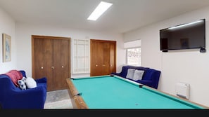 Ein Zimmer mit Billardtisch, zwei blauen Sofas, Holzschränken, einem Fernseher und einem Fenster mit Aussicht.