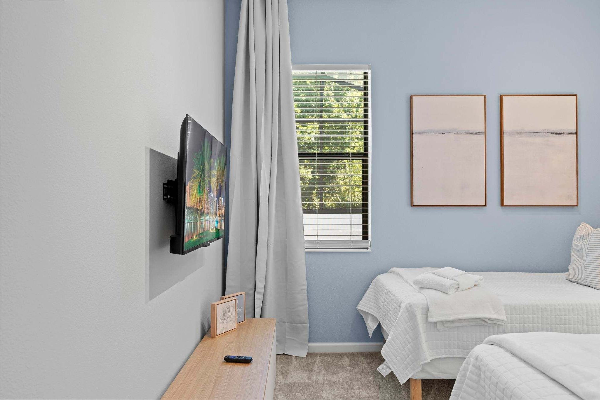 Un dormitorio con televisión, dos camas, una ventana con persianas y cuadros en la pared.