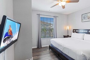Une chambre d’hôtel avec un lit, un téléviseur fixé au mur, un ventilateur de plafond, une fenêtre avec des rideaux et une table de chevet avec une lampe.