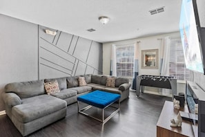 Een moderne woonkamer met een modulaire bank, een blauwe poef en een tafelvoetbaltafel.