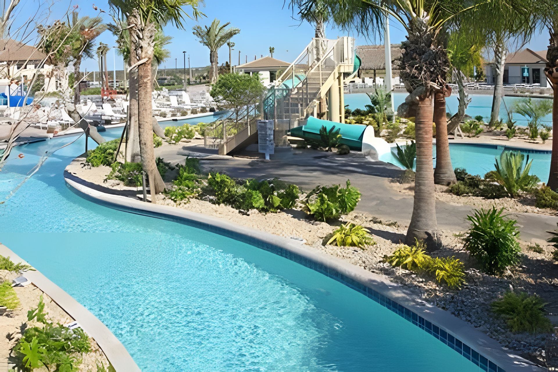 A swimming pool with a curved design, surrounded by palm trees and a small island. There are lounge chairs and a water slide in the background.