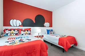 Ein Schlafzimmer mit zwei Betten, beide mit roter Bettdecke mit Zeichentrickfiguren. Darüber befindet sich eine große Wandmalerei, und ein weißer Nachttisch mit einer Lampe.