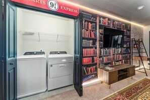 A laundry room with two white washers and dryers, a bookshelf filled with books, a television, and a wooden coffee table. - Themed 5BR Home w/ Pool · Windsor Island Resort (Davenport)
