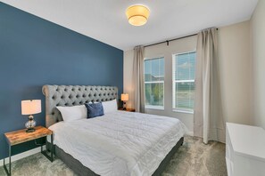 A bedroom with a tufted headboard, a white bedspread, a nightstand with a lamp, and a window with curtains. - Storey Lake 6BR Home w/ Pool & Game Room (Kissimmee)